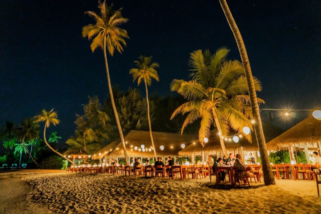 Magical beachfront dining under palm trees and string lights during Easter 2026 at Sun Siyam Resorts, Maldives. Guests enjoying a special dinner on the white sandy beach with thatched-roof restaurants in the background, creating a romantic and festive atmosphere at Easter 2026 Sun Siyam Maldives.