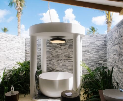 Open-air bathroom in a Beach Residence at Joy Island Maldives featuring a freestanding white bathtub under a circular canopy, stone walls, and tropical plants under blue sky