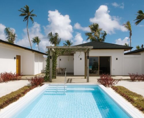 Private rectangular pool of a Beach Residence at Joy Island Maldives facing the villa buildings, wooden pergola, and palm trees under a bright blue sky