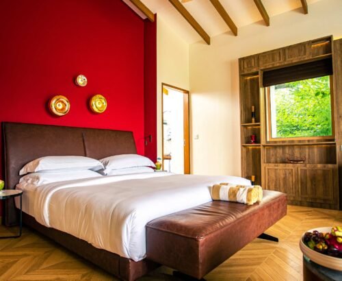 Beach Suite bedroom at Joy Island Maldives featuring a large bed against a vibrant red accent wall with gold circular lights, wooden flooring, and tropical greenery outside the window