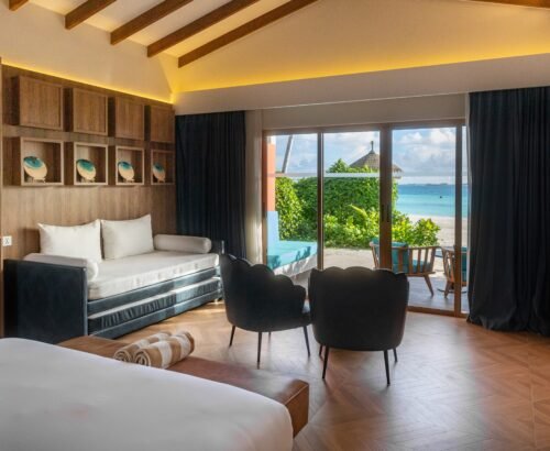 Spacious Beach Suite interior at Joy Island Maldives featuring a large bed, daybed, modern seating, and open glass doors leading to the beach and turquoise lagoon