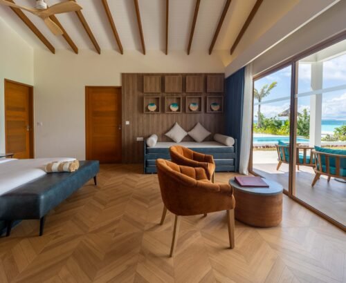 Interior of a Beach Suite at Joy Island Maldives showing a sofa, modern chairs, wooden flooring, and large glass doors opening to the private pool and tropical garden