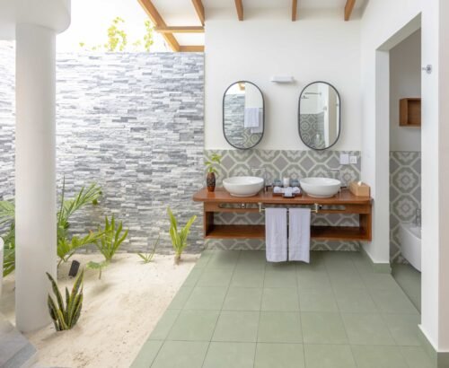 Open-air bathroom in a Beach Suite at Joy Island Maldives featuring dual white vessel sinks on a wooden vanity, oval mirrors, and a tropical outdoor bathing area with stone wall