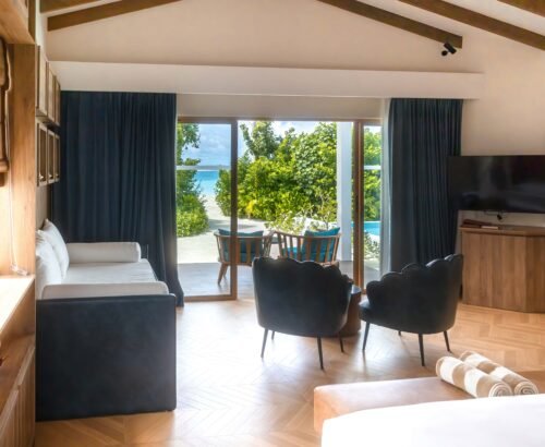 Spacious Beach Suite interior at Joy Island Maldives with sofa, modern chairs, and large glass doors opening to outdoor seating and private pool area