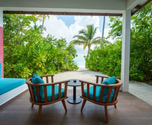 Covered outdoor seating area of a Beach Suite at Joy Island Maldives looking through tropical greenery toward the white sand beach and turquoise lagoon