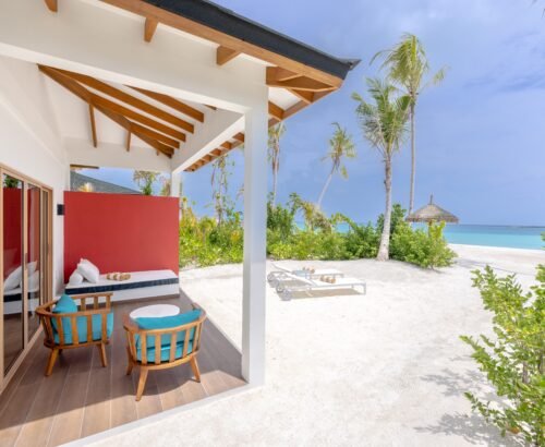 Covered patio of a Beach Suite at Joy Island Maldives with turquoise seating and daybed overlooking the white sand beach and turquoise lagoon