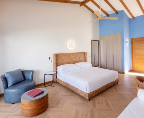 Spacious Beach Villa bedroom at Joy Island Maldives featuring a large bed with brown headboard, blue accent wall, wooden flooring, and modern seating area