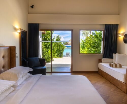 Bright Beach Villa bedroom at Joy Island Maldives with large bed, daybed, and open glass doors framing views of tropical greenery, white sand, and turquoise lagoon