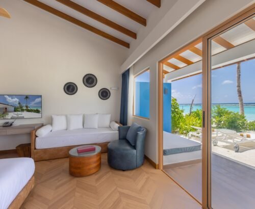 Wake up to natural light in a Beach Villa bedroom at Joy Island Maldives, where a comfortable bed and daybed face large glass doors that open toward the white sand and turquoise lagoon. Soft curtains, warm wood tones, and clean design create a calm and inviting space that connects the indoors with the tropical surroundings. Guests can step straight from the room onto the sandy path leading to the beach. Our Joy Island Maldives tour package includes stays in Beach Villas with this open layout, offering a peaceful and scenic base in the North Malé Atoll. Book a Joy Island Maldives tour package and enjoy mornings that begin with ocean views right from your Beach Villa bedroom.