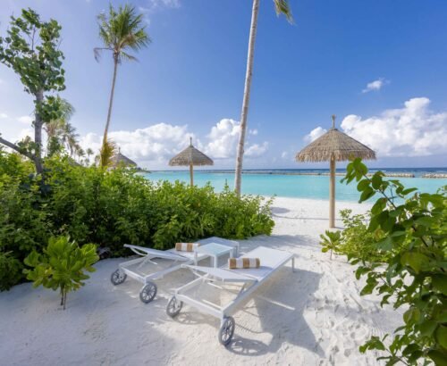 White sun loungers and thatched umbrellas on the soft sand in front of a Beach Villa at Joy Island Maldives overlooking the turquoise lagoon and blue sky