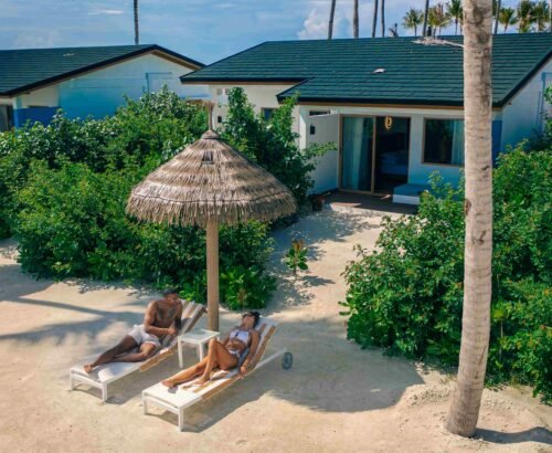 Couple relaxing on sun loungers under a thatched umbrella in front of a Beach Villa at Joy Island Maldives surrounded by tropical greenery and palm trees