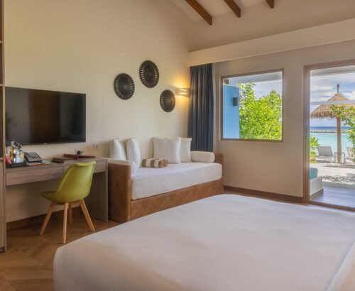 Family Beach Villa interior at Joy Island Maldives with a large bed, daybed sofa, desk area, and open sliding doors leading to the turquoise lagoon and beach