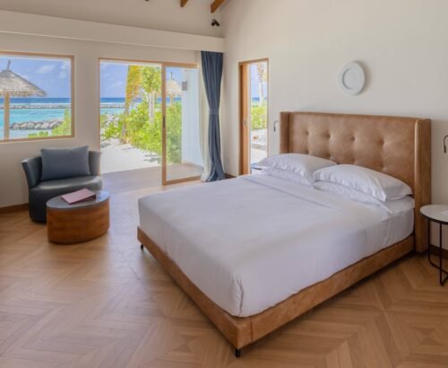 Bright Family Beach Villa bedroom at Joy Island Maldives with a large bed, seating area, and open glass doors framing views of the turquoise lagoon and beach