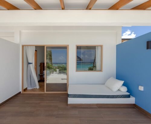 Covered outdoor area of a Family Beach Villa at Joy Island Maldives with a built-in daybed against a blue wall and views of the lagoon through the window