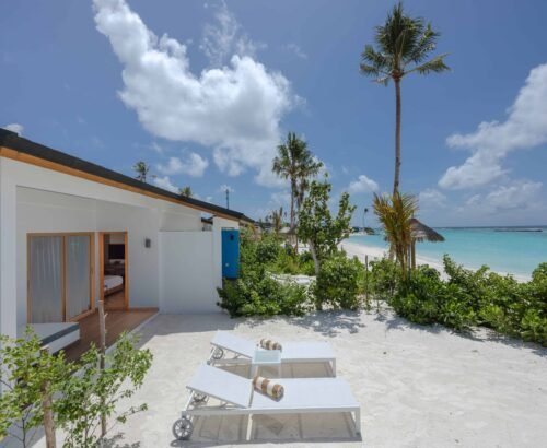 Family Beach Villa at Joy Island Maldives with white sun loungers on the soft sand overlooking the turquoise lagoon and palm trees under a blue sky