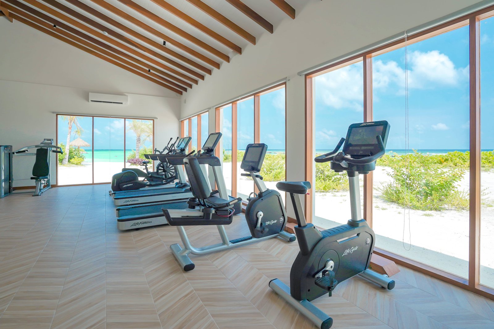 Modern fitness centre at Joy Island Maldives with treadmills and exercise bikes facing large windows overlooking the white sand beach and turquoise lagoon