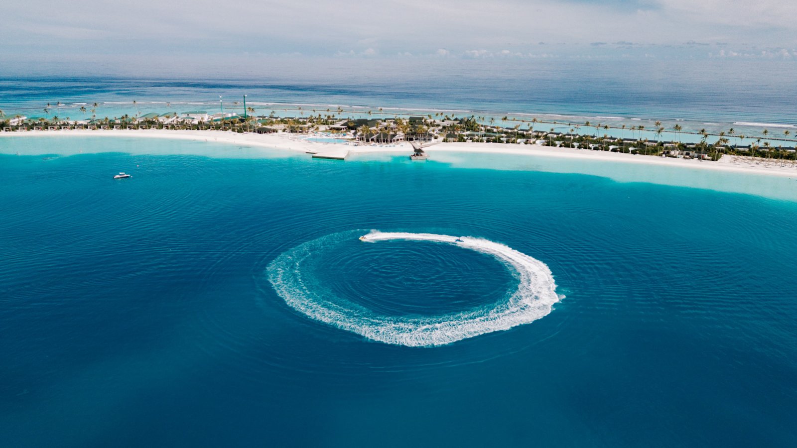 Aerial view of a jet ski creating a circular white wake in the clear turquoise lagoon at Joy Island Maldives with the full island resort in the background