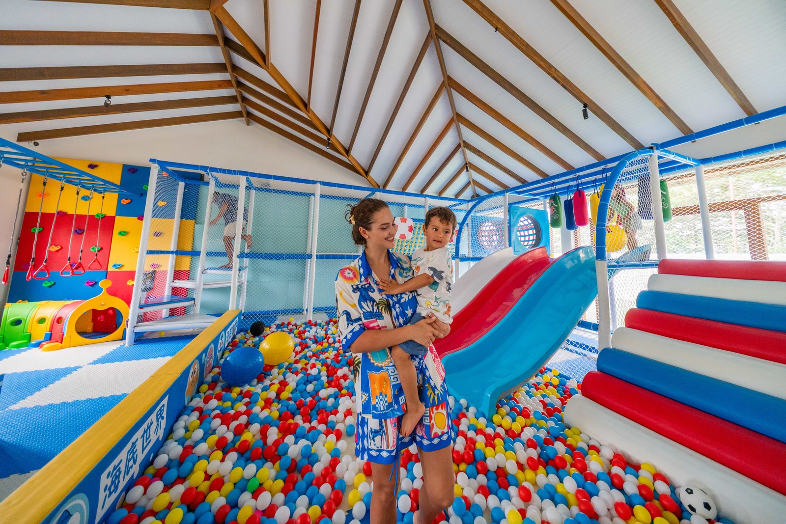 Mother holding young child inside the colorful Kiddie Cove kids club at Joy Island Maldives with ball pit, slides and soft play equipment