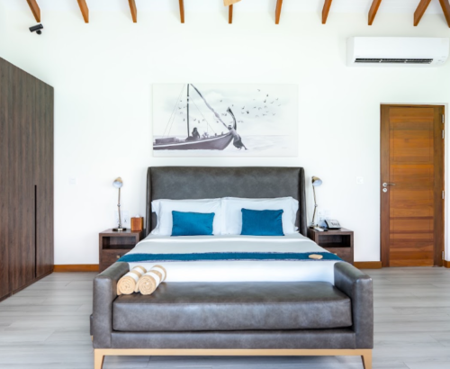 Modern Lagoon Residence bedroom at Joy Island Maldives featuring a large bed with grey headboard, teal pillows, wooden wardrobe, and a sailboat artwork on the wall