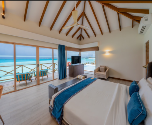 Spacious Lagoon Residence interior at Joy Island Maldives with a large bed, teal accents, high wooden ceiling, and open glass doors leading to the overwater deck and turquoise lagoon