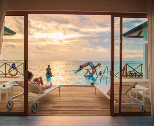View from inside a Lagoon Residence at Joy Island Maldives looking out through open glass doors to the private pool where guests dive and relax under a soft sunset sky