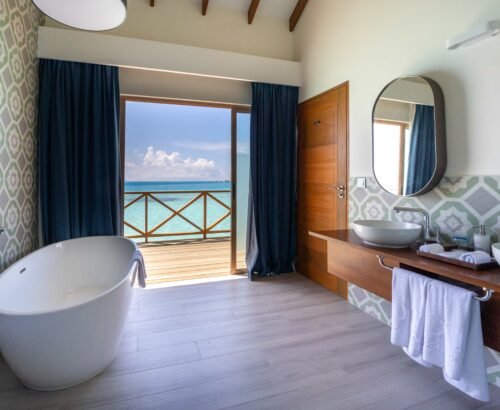 Lagoon Suite bathroom at Joy Island Maldives featuring a freestanding white bathtub, dual vessel sinks, and open glass doors looking out to the turquoise lagoon