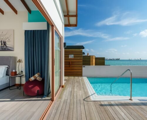 Lagoon Suite interior at Joy Island Maldives with a bedroom opening through glass doors to a private pool and panoramic turquoise lagoon views under a blue sky