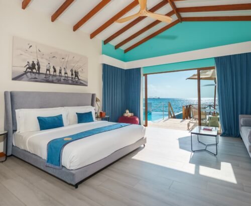 Bright Lagoon Suite bedroom at Joy Island Maldives with a large bed, teal accents, and open glass doors leading to a private pool and turquoise lagoon under a blue sky