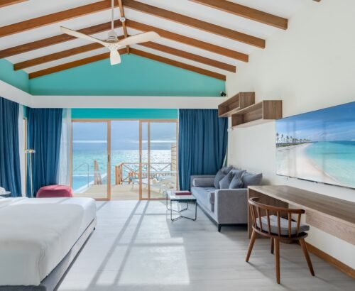 Spacious Lagoon Suite interior at Joy Island Maldives with a bed, grey sofa, desk area, and large glass doors opening to a private pool and turquoise lagoon under a bright sky