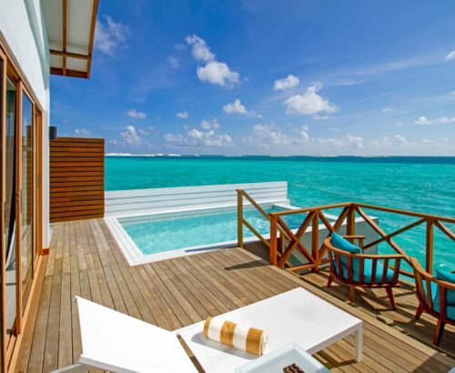 Overwater deck of a Lagoon Suite at Joy Island Maldives with a private rectangular pool, turquoise seating, and panoramic views of the clear turquoise lagoon under a blue sky