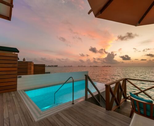 Private pool on the overwater deck of a Lagoon Suite at Joy Island Maldives glowing under a colorful sunset sky with pink and orange clouds over the lagoon