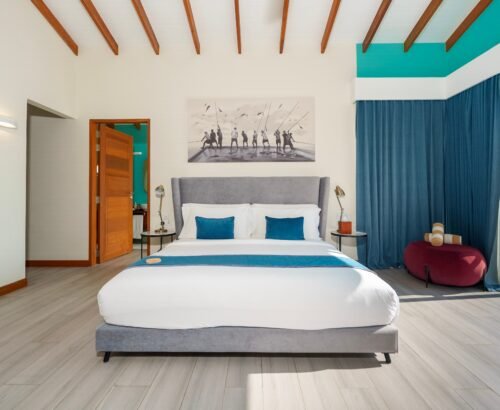 Spacious Lagoon Suite bedroom at Joy Island Maldives featuring a large bed with grey headboard, teal accents, wooden flooring, and glass doors opening to the private pool and lagoon