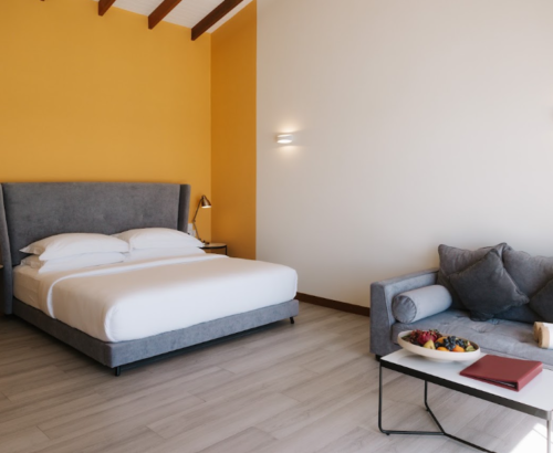 Bright Lagoon Villa bedroom at Joy Island Maldives featuring a large bed against a yellow accent wall, grey sofa, and modern seating area with light wood flooring