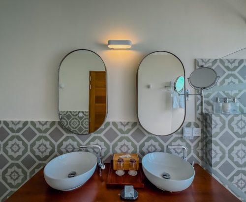 Bathroom vanity in a Lagoon Villa at Joy Island Maldives featuring dual white vessel sinks on a wooden counter, oval mirrors, and patterned grey and white tiles