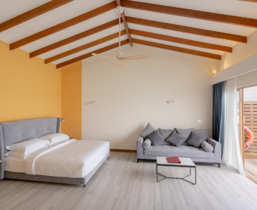 Bright Lagoon Villa interior at Joy Island Maldives featuring a large bed, grey sofa, high wooden ceiling, and open glass doors leading to an overwater wooden deck with ocean views