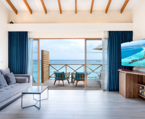 Lagoon Villa living space at Joy Island Maldives with a grey sofa, large glass doors opening to the overwater deck, and panoramic views of the turquoise lagoon