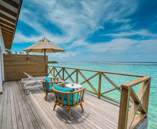 Wooden overwater deck of a Lagoon Villa at Joy Island Maldives with turquoise cushioned chairs, sun loungers, and panoramic views of the clear turquoise lagoon under a blue sky