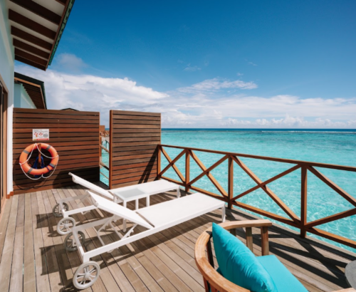 Wooden overwater deck of a Lagoon Villa at Joy Island Maldives with white sun loungers, turquoise seating, and panoramic views of the turquoise lagoon under a clear blue sky