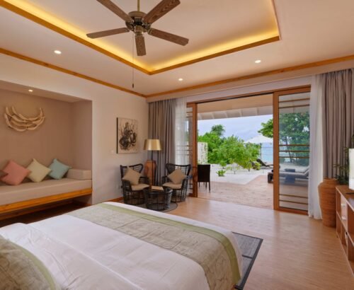 Spacious Beach Villa bedroom at Brennia Kottefaru Maldives with a large bed, seating area, and open sliding doors leading to a private deck, sun loungers, and lagoon views