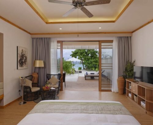 Spacious Beach Villa bedroom at Brennia Kottefaru Maldives with a large bed, seating area, and open sliding doors leading to a beachfront deck with sun loungers and lagoon views