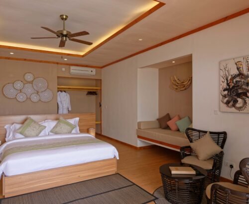 Spacious Beach Villa bedroom at Brennia Kottefaru Maldives with a large wooden bed, open wardrobe, built-in daybed, and rattan seating area under warm lighting