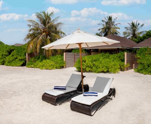 Two white sun loungers under a large umbrella on the soft sand outside a Beach Villa at Brennia Kottefaru Maldives with tropical plants and villa buildings in the background