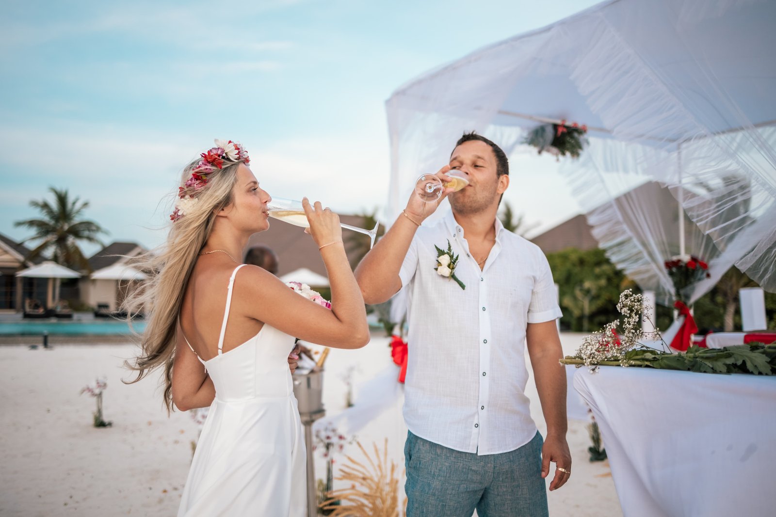 Couple toasting with champagne during a beach wedding at Brennia Kottefaru Maldives, with a white canopy, floral décor, and soft sand under a clear sky