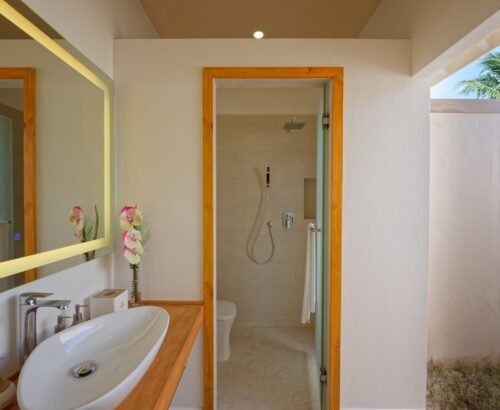Modern bathroom in a Bougain Villa at Brennia Kottefaru Maldives featuring a white vessel sink, illuminated wooden-framed mirror, and open shower area with palm tree views