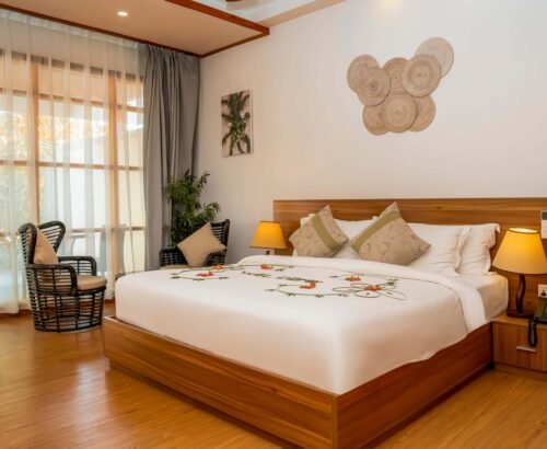 Elegant Bougain Villa bedroom at Brennia Kottefaru Maldives featuring a large wooden bed with flower petal decoration, soft lighting, and rattan seating near the window