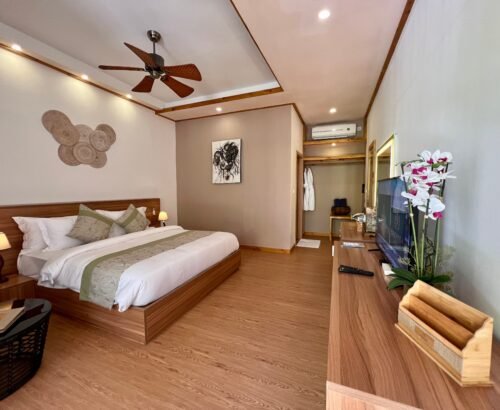 Spacious Bougain Villa bedroom at Brennia Kottefaru Maldives with a large wooden bed, green accents, TV console with orchids, and open wardrobe area