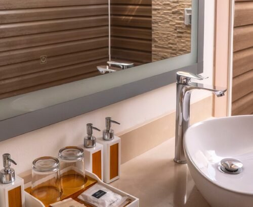 Close-up of a Deluxe Beach Pool Villa bathroom vanity at Brennia Kottefaru Maldives with a white vessel sink, chrome faucet, toiletries, and a large mirror reflecting the outdoor shower