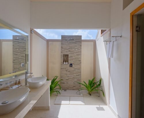 Deluxe Beach Pool Villa bathroom at Brennia Kottefaru Maldives featuring dual white vessel sinks on a marble vanity and an open stone-tiled outdoor shower with tropical plants and natural light