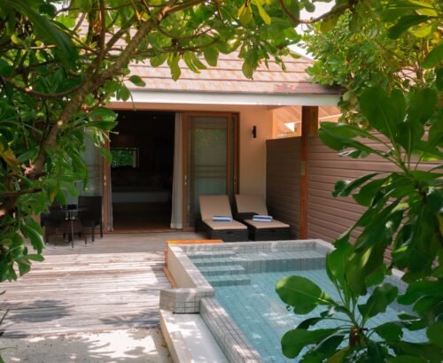 Deluxe Beach Pool Villa at Brennia Kottefaru Maldives framed by tropical greenery with a private blue pool, wooden deck, sun loungers, and open sliding doors leading into the villa