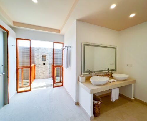 Deluxe Water Pool Villa bathroom at Brennia Kottefaru Maldives featuring dual white vessel sinks on a clean vanity and an open stone-tiled outdoor shower framed by wooden panels